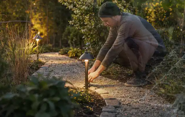 Person richtet eine niedrige warm leuchtende Gartenlampe am Kiesweg aus; Thema Mediterraner Garten, Schwerpunkt Kosten; Praxisalltag.