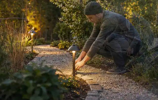 Person richtet eine niedrige warm leuchtende Gartenlampe am Kiesweg aus; Thema Mediterraner Garten, Schwerpunkt Naturgarten; Kontrolle.