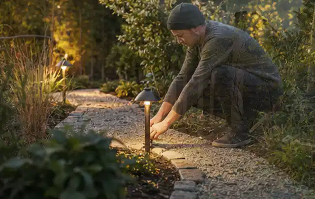Person richtet eine niedrige warm leuchtende Gartenlampe am Kiesweg aus; Thema Metallzäune, Schwerpunkt Aufbau; Ausgangslage.