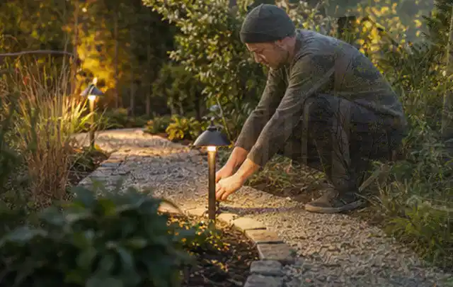 Person richtet eine niedrige warm leuchtende Gartenlampe am Kiesweg aus; Thema Metallzäune, Schwerpunkt Checkliste; Praxisalltag.