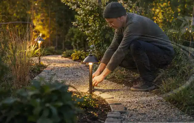 Person richtet eine niedrige warm leuchtende Gartenlampe am Kiesweg aus; Thema Metallzäune, Schwerpunkt Dimensionierung; Umsetzung.