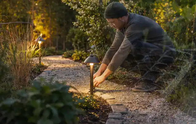 Person richtet eine niedrige warm leuchtende Gartenlampe am Kiesweg aus; Thema Metallzäune, Schwerpunkt Gärten mit kleiner Fläche; Ergebnis.