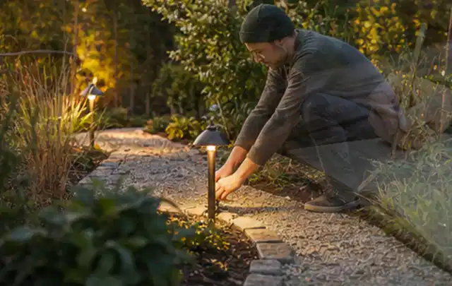 Person richtet eine niedrige warm leuchtende Gartenlampe am Kiesweg aus; Thema Metallzäune, Schwerpunkt Gestaltung; Ausgangslage.