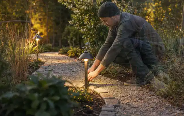 Person richtet eine niedrige warm leuchtende Gartenlampe am Kiesweg aus; Thema Metallzäune, Schwerpunkt Materialwahl; Ausgangslage.