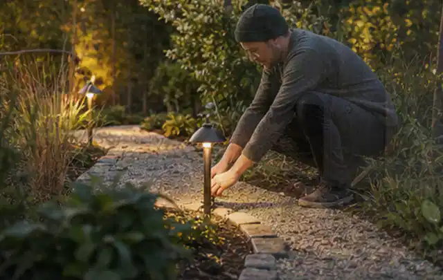 Person richtet eine niedrige warm leuchtende Gartenlampe am Kiesweg aus; Thema Metallzäune, Schwerpunkt Sichtschutz; Umsetzung.