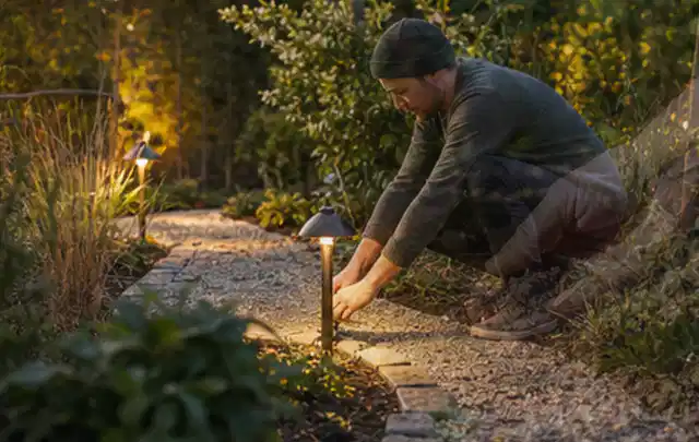 Person richtet eine niedrige warm leuchtende Gartenlampe am Kiesweg aus; Thema Metallzäune, Schwerpunkt Standort; Kontrolle.