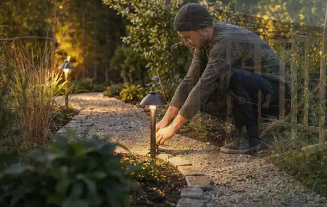 Person richtet eine niedrige warm leuchtende Gartenlampe am Kiesweg aus; Thema Mobiler Sichtschutz, Schwerpunkt Checkliste; Praxisalltag.
