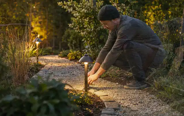Person richtet eine niedrige warm leuchtende Gartenlampe am Kiesweg aus; Thema Mobiler Sichtschutz, Schwerpunkt Fehlerhilfe; Kontrolle.