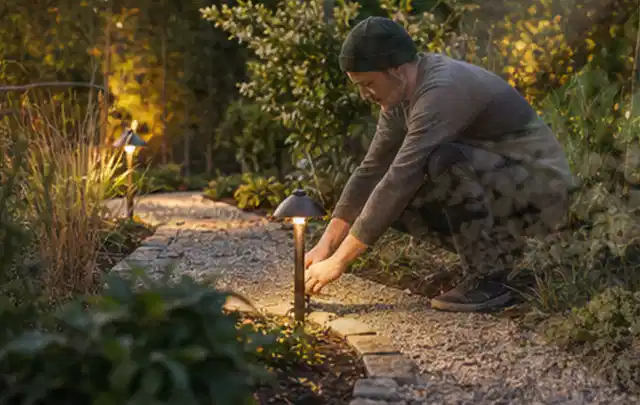 Person richtet eine niedrige warm leuchtende Gartenlampe am Kiesweg aus; Thema Mobiler Sichtschutz, Schwerpunkt Fundament; Umsetzung.
