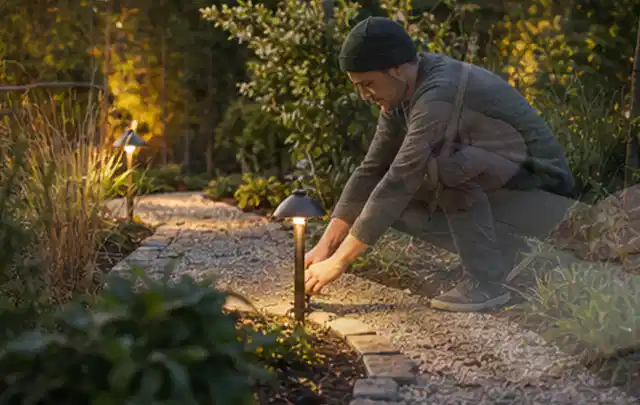 Person richtet eine niedrige warm leuchtende Gartenlampe am Kiesweg aus; Thema Mobiler Sichtschutz, Schwerpunkt Gestaltung; Umsetzung.