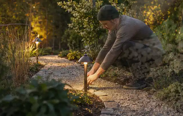 Person richtet eine niedrige warm leuchtende Gartenlampe am Kiesweg aus; Thema Mobiler Sichtschutz, Schwerpunkt Pflege; Praxisalltag.