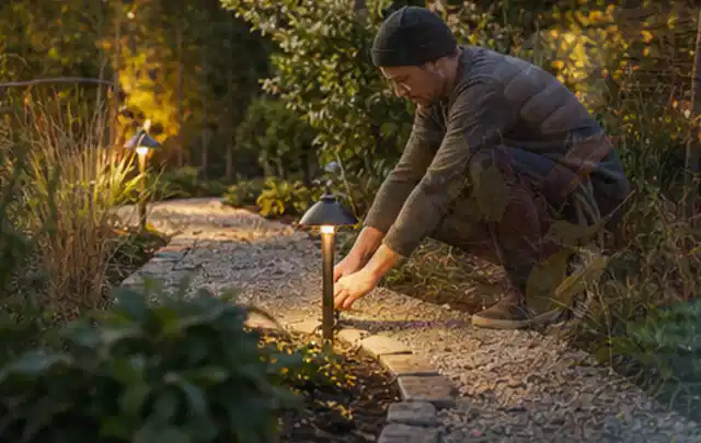 Person richtet eine niedrige warm leuchtende Gartenlampe am Kiesweg aus; Thema Moderner Garten, Schwerpunkt Materialwahl; Kontrolle.