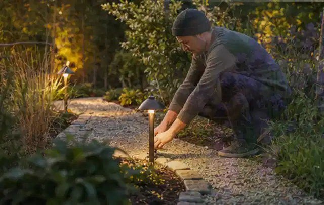 Person richtet eine niedrige warm leuchtende Gartenlampe am Kiesweg aus; Thema Moderner Garten, Schwerpunkt Planung; Kontrolle.