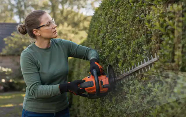 Person schneidet eine dichte Gartenhecke mit Schutzbrille und sicherem Stand; Thema Motorhacken, Schwerpunkt Dimensionierung; Umsetzung.