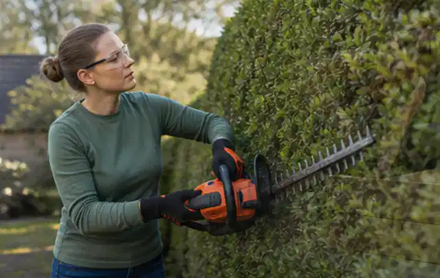Person schneidet eine dichte Gartenhecke mit Schutzbrille und sicherem Stand; Thema Motorsensen, Schwerpunkt Checkliste; Kontrolle.