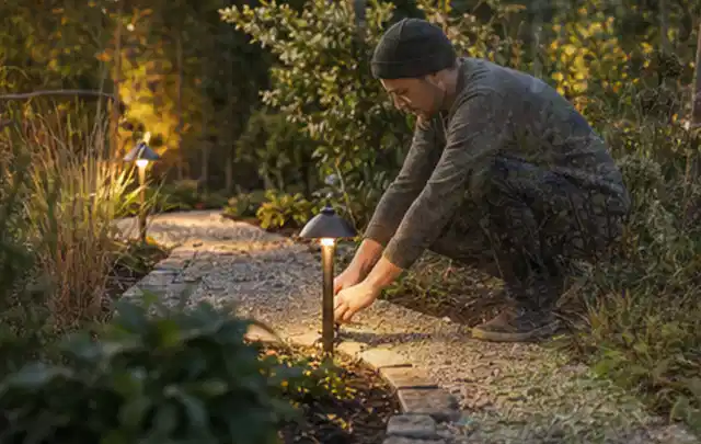 Person richtet eine niedrige warm leuchtende Gartenlampe am Kiesweg aus; Thema Naturgarten, Schwerpunkt Anleitung; Praxisalltag.