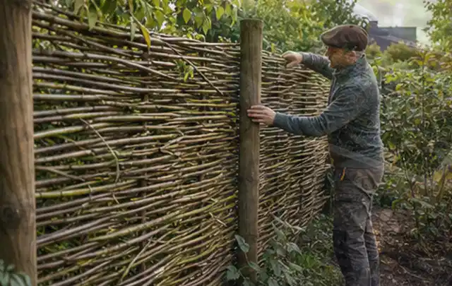 Person befestigt geflochtene Weidenruten an stabilen Holzpfosten im Garten; Thema Naturgarten, Schwerpunkt Checkliste; Praxisalltag.