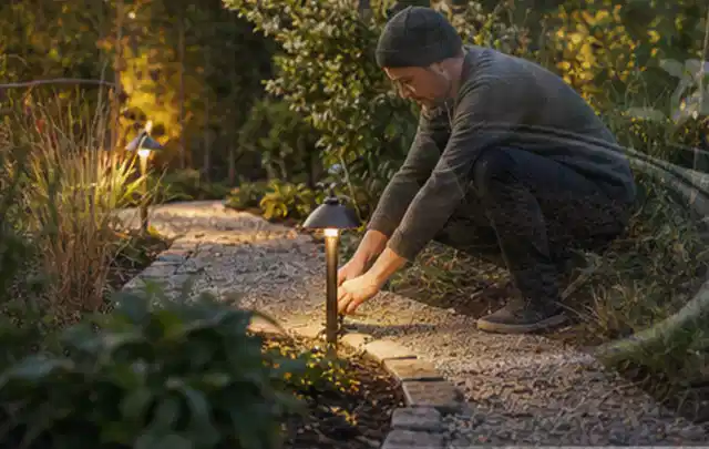 Person richtet eine niedrige warm leuchtende Gartenlampe am Kiesweg aus; Thema Naturgarten, Schwerpunkt Gärten mit kleiner Fläche; Ergebnis.