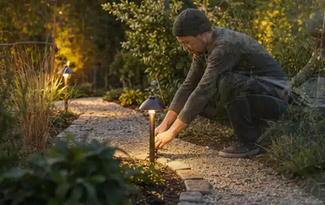 Person richtet eine niedrige warm leuchtende Gartenlampe am Kiesweg aus; Thema Naturgarten, Schwerpunkt Hanglage; Umsetzung.