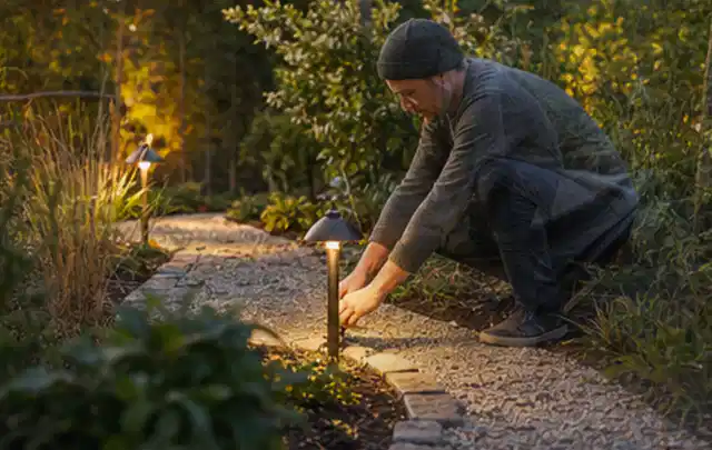 Person richtet eine niedrige warm leuchtende Gartenlampe am Kiesweg aus; Thema Naturgarten, Schwerpunkt Privatsphäre; Ausgangslage.