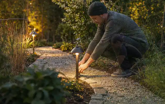Person richtet eine niedrige warm leuchtende Gartenlampe am Kiesweg aus; Thema Outdoor Küchen, Schwerpunkt Ersatzteile; Ergebnis.