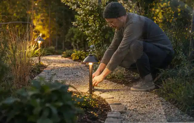 Person richtet eine niedrige warm leuchtende Gartenlampe am Kiesweg aus; Thema Outdoor Küchen, Schwerpunkt Fehlerhilfe; Ergebnis.