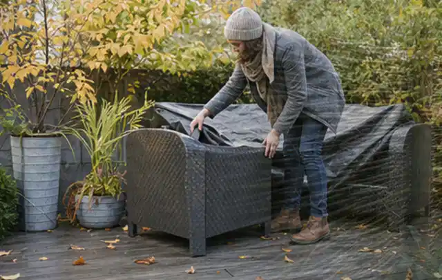 Person deckt Gartenmöbel vor der kalten Jahreszeit sorgfältig ab; Thema Outdoor Küchen, Schwerpunkt Gärten mit kleiner Fläche; Praxisalltag.
