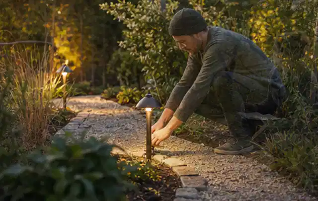 Person richtet eine niedrige warm leuchtende Gartenlampe am Kiesweg aus; Thema Outdoor Küchen, Schwerpunkt Nachhaltigkeit; Umsetzung.