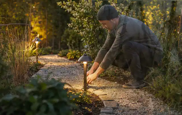 Person richtet eine niedrige warm leuchtende Gartenlampe am Kiesweg aus; Thema Outdoor Küchen, Schwerpunkt Outdoor Freizeit; Umsetzung.