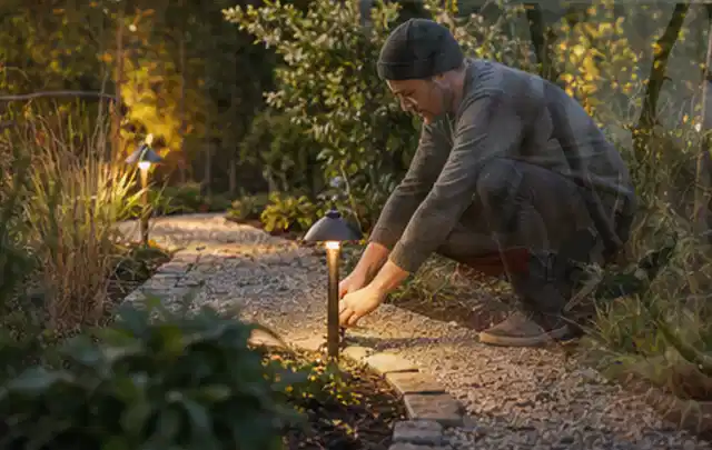 Person richtet eine niedrige warm leuchtende Gartenlampe am Kiesweg aus; Thema Outdoor Küchen, Schwerpunkt Winterlager; Ergebnis.