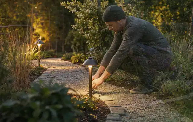 Person richtet eine niedrige warm leuchtende Gartenlampe am Kiesweg aus; Thema Pelletgrills, Schwerpunkt Anwendung; Praxisalltag.
