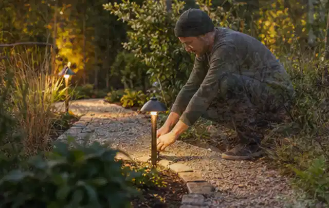 Person richtet eine niedrige warm leuchtende Gartenlampe am Kiesweg aus; Thema Pelletgrills, Schwerpunkt Dimensionierung; Kontrolle.