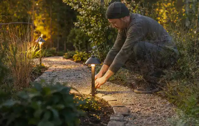 Person richtet eine niedrige warm leuchtende Gartenlampe am Kiesweg aus; Thema Pelletgrills, Schwerpunkt Fehlerhilfe; Praxisalltag.
