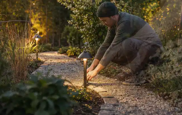 Person richtet eine niedrige warm leuchtende Gartenlampe am Kiesweg aus; Thema Pelletgrills, Schwerpunkt Leistung; Umsetzung.