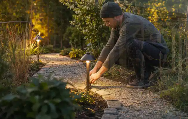 Person richtet eine niedrige warm leuchtende Gartenlampe am Kiesweg aus; Thema Pelletgrills, Schwerpunkt Outdoor Freizeit; Kontrolle.
