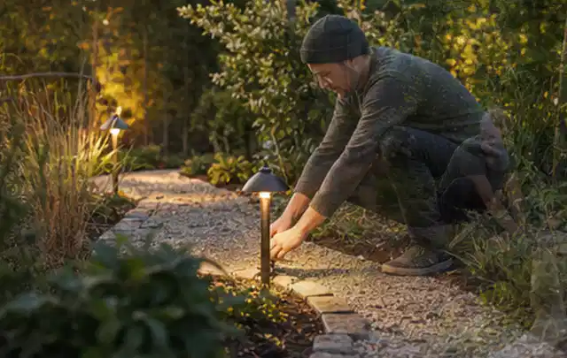 Person richtet eine niedrige warm leuchtende Gartenlampe am Kiesweg aus; Thema Pelletgrills, Schwerpunkt Wartung; Kontrolle.