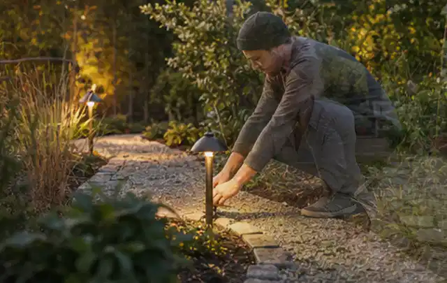 Person richtet eine niedrige warm leuchtende Gartenlampe am Kiesweg aus; Thema Pergolen, Schwerpunkt Checkliste; Praxisalltag.