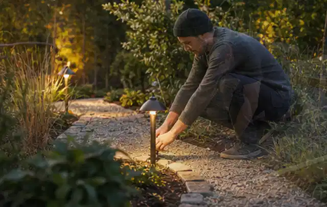 Person richtet eine niedrige warm leuchtende Gartenlampe am Kiesweg aus; Thema Pergolen, Schwerpunkt Ersatzteile; Ausgangslage.