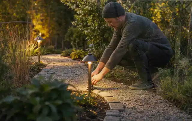 Person richtet eine niedrige warm leuchtende Gartenlampe am Kiesweg aus; Thema Pergolen, Schwerpunkt Garten gestalten; Ergebnis.