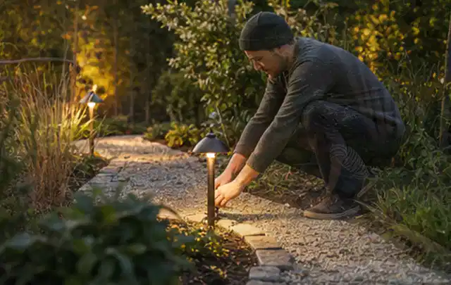 Person richtet eine niedrige warm leuchtende Gartenlampe am Kiesweg aus; Thema Pergolen, Schwerpunkt Kosten; Ergebnis.
