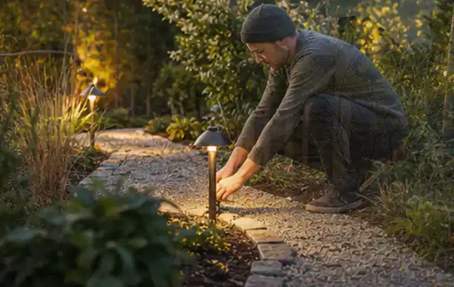 Person richtet eine niedrige warm leuchtende Gartenlampe am Kiesweg aus; Thema Pergolen, Schwerpunkt Outdoor Freizeit; Ergebnis.