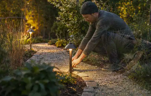 Person richtet eine niedrige warm leuchtende Gartenlampe am Kiesweg aus; Thema Pergolen, Schwerpunkt Reparatur; Kontrolle.