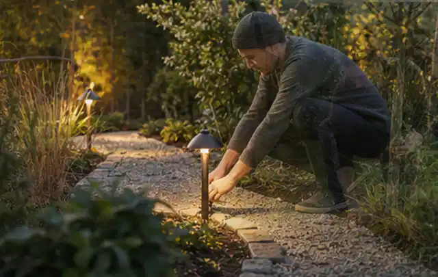 Person richtet eine niedrige warm leuchtende Gartenlampe am Kiesweg aus; Thema Pergolen, Schwerpunkt Testkriterien; Umsetzung.