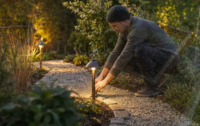 Person richtet eine niedrige warm leuchtende Gartenlampe am Kiesweg aus; Thema Pergolen, Schwerpunkt Vergleich; Ergebnis.