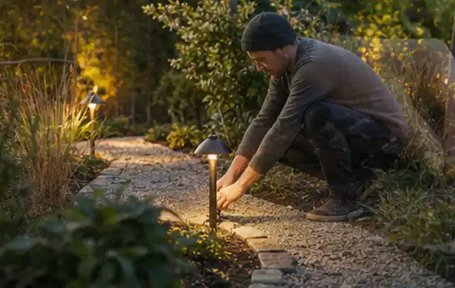 Person richtet eine niedrige warm leuchtende Gartenlampe am Kiesweg aus; Thema Pergolen, Schwerpunkt Wartung; Kontrolle.