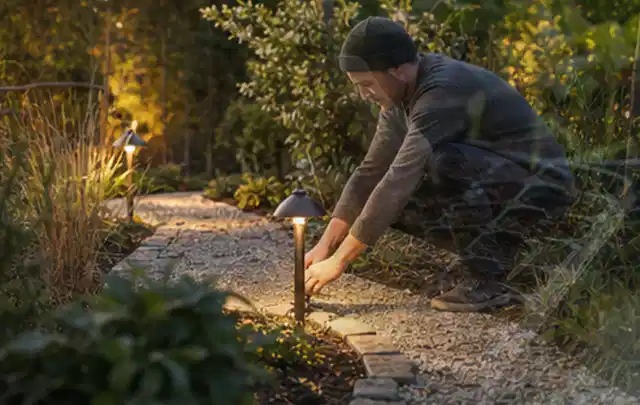 Person richtet eine niedrige warm leuchtende Gartenlampe am Kiesweg aus; Thema Pergolen, Schwerpunkt Winterlager; Ausgangslage.