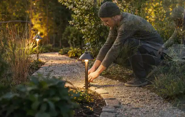 Person richtet eine niedrige warm leuchtende Gartenlampe am Kiesweg aus; Thema Pflegeleichter Garten, Schwerpunkt Gärten mit kleiner Fläche; Kontrolle.