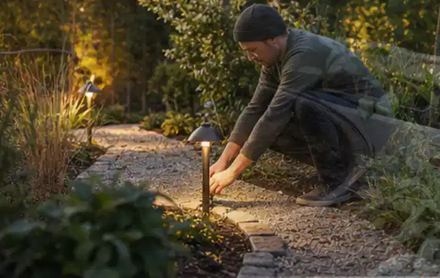 Person richtet eine niedrige warm leuchtende Gartenlampe am Kiesweg aus; Thema Pflegeleichter Garten, Schwerpunkt Materialwahl; Ausgangslage.