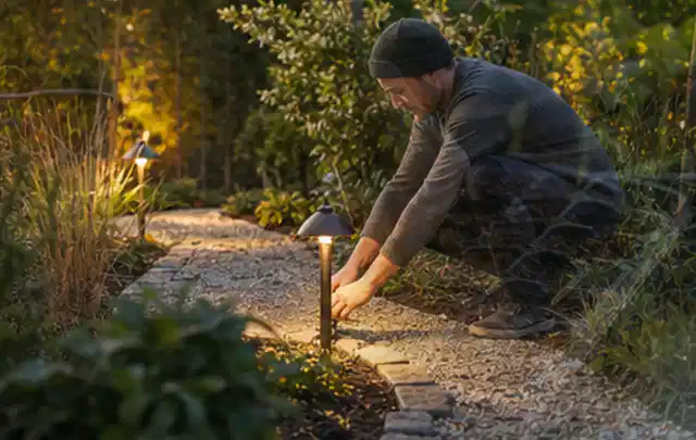 Person richtet eine niedrige warm leuchtende Gartenlampe am Kiesweg aus; Thema Pflegeleichter Garten, Schwerpunkt Planung; Praxisalltag.