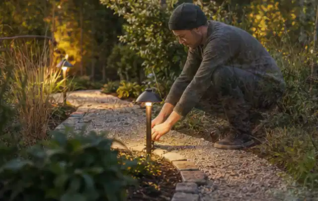 Person richtet eine niedrige warm leuchtende Gartenlampe am Kiesweg aus; Thema Pflegeleichter Garten, Schwerpunkt Privatsphäre; Praxisalltag.